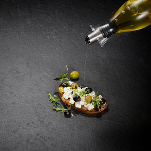A drizzle of olive oil from the Cole & Mason UK Haverhill Oil & Vinegar Pourers Set 450ml enhances bread with feta, olives, and arugula, all beautifully arranged on a dark textured surface.