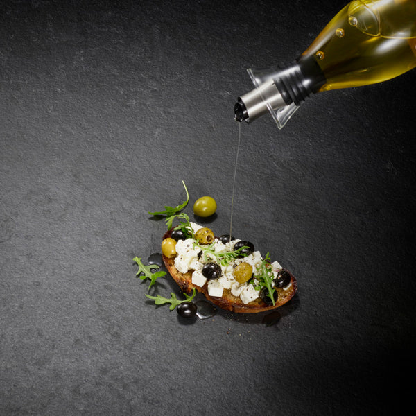 A drizzle of olive oil from the Cole & Mason UK Haverhill Oil & Vinegar Pourers Set 450ml enhances bread with feta, olives, and arugula, all beautifully arranged on a dark textured surface.