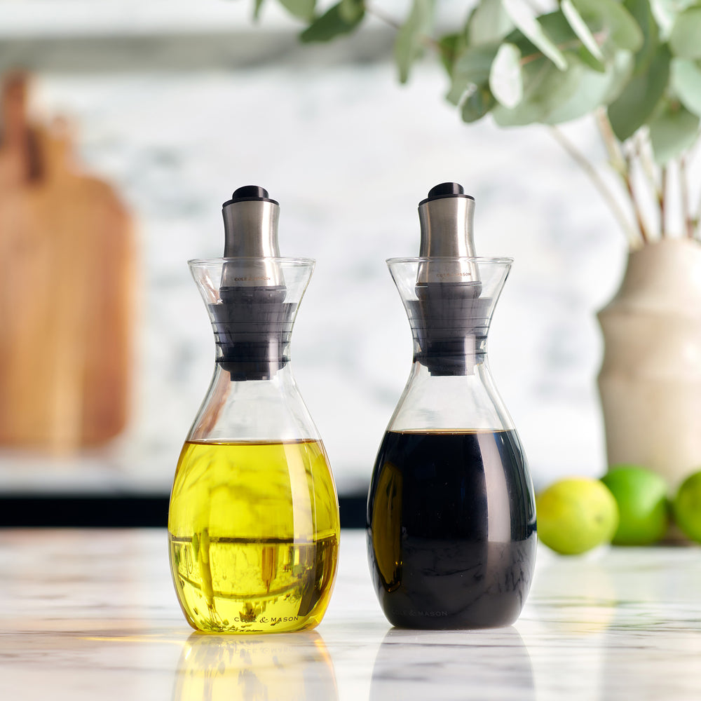 The Cole & Mason UK Haverhill Oil & Vinegar Pourers Set 450ml includes two glass pourers with stainless steel spouts, adjustable rubber stoppers, and a drip return system—one holds yellow oil, the other dark brown vinegar.