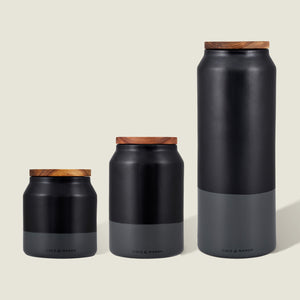 The Cole & Mason Hinxton Ceramic Storage Jar Set features three black and gray jars of varying heights with airtight wooden lids, arranged on a light beige background—a modern, minimalist choice for elegant kitchen storage.