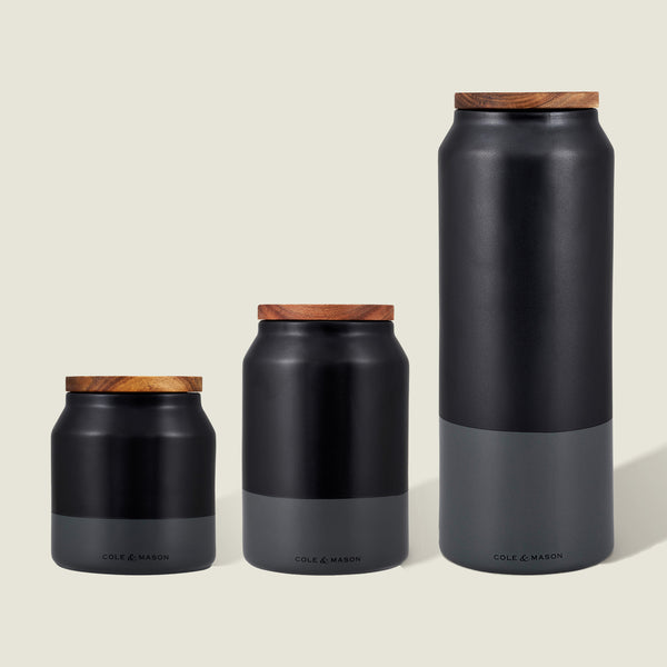 The Cole & Mason Hinxton Ceramic Storage Jar Set features three black and gray jars of varying heights with airtight wooden lids, arranged on a light beige background—a modern, minimalist choice for elegant kitchen storage.
