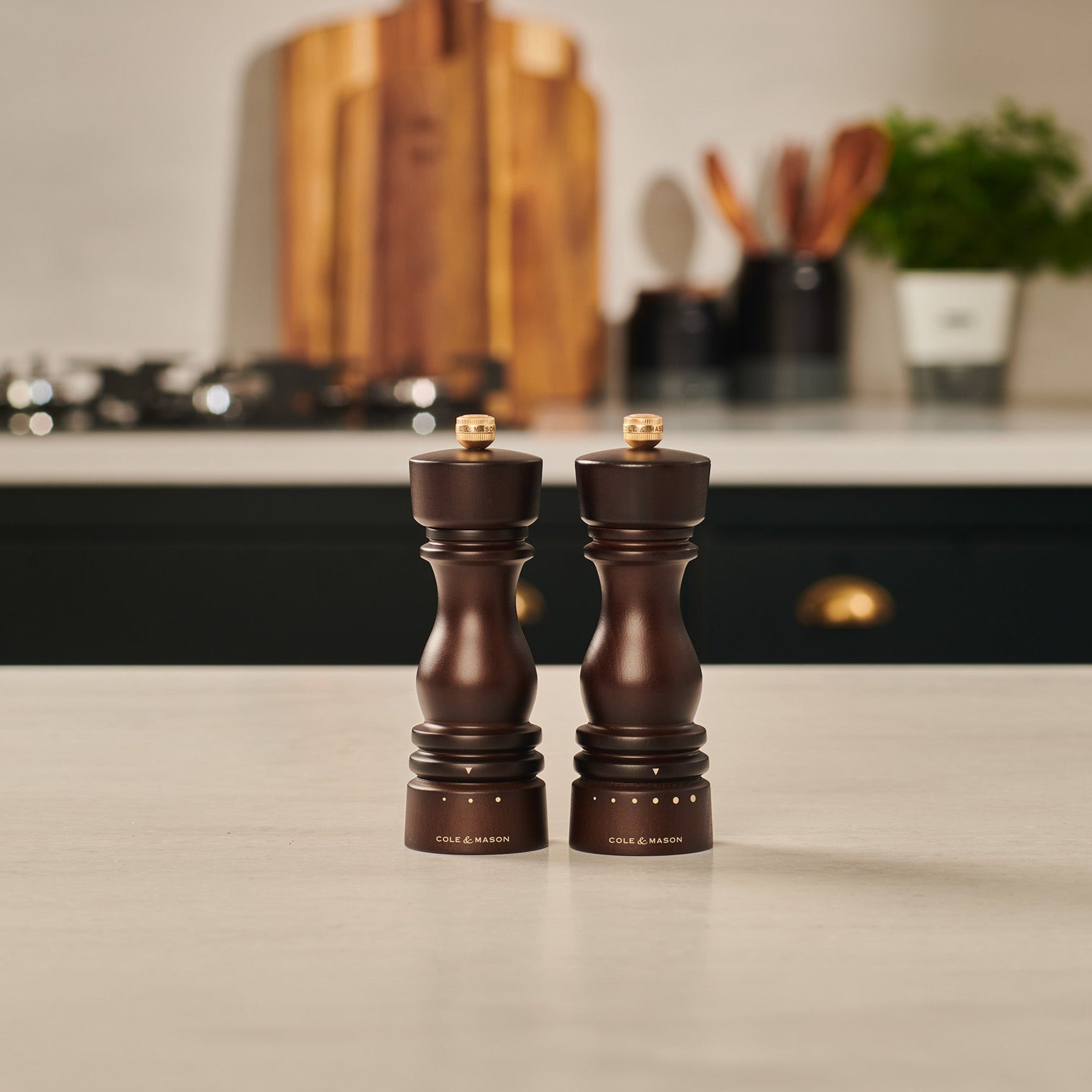 Two dark wooden pepper and salt grinders stand side by side on a light kitchen countertop, with a blurred modern kitchen and wooden utensils in the background.
