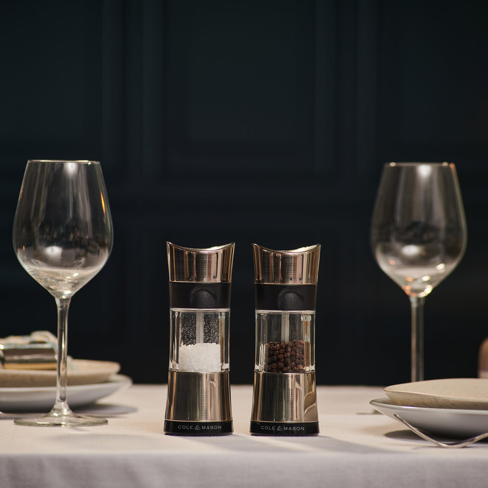 Two Cole & Mason Horsham Salt & Pepper Mills are shown side by side; the left holds coarse salt, the right, whole peppercorns. Both have clear midsections to display contents and offer high-performance grinding with adjustable settings.
