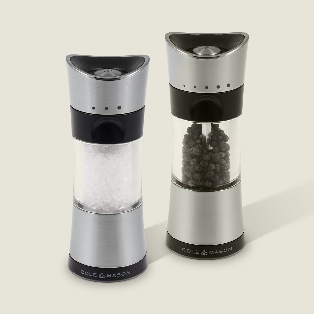 Two Cole & Mason Horsham Salt & Pepper Mills are shown side by side; the left holds coarse salt, the right, whole peppercorns. Both have clear midsections to display contents and offer high-performance grinding with adjustable settings.