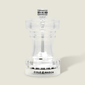 Showcased against a light beige background, the Cole & Mason Acrylic Salt Shaker & Pepper Mill Set (104 mm) features a clear acrylic body with a silver knob on top and “COLE & MASON” inscribed in black on the base; this elegant pepper mill offers an adjustable grinder for customized seasoning.