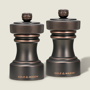Two Cole & Mason Hoxton Salt and Pepper Mills in antique bronze, each with a dark finish, textured knurled tops, and the Cole & Mason brand name engraved in gold at the base, stand side by side against a plain light background. This sophisticated set features a precision grinding system for effortless seasoning every time.