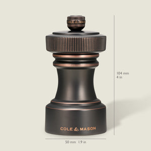 A dark metallic pepper grinder from the Cole & Mason Hoxton Salt and Pepper Mill Set stands upright, showcasing an antique bronze finish. Dimensions are indicated as 104 mm (4 in) tall and 50 mm (1.9 in) wide, set against a light beige background.