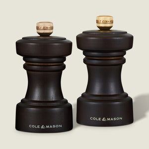 Two dark wood Cole & Mason mills, one for pepper and one for salt, with gold knobs on top, placed side by side on a light background.