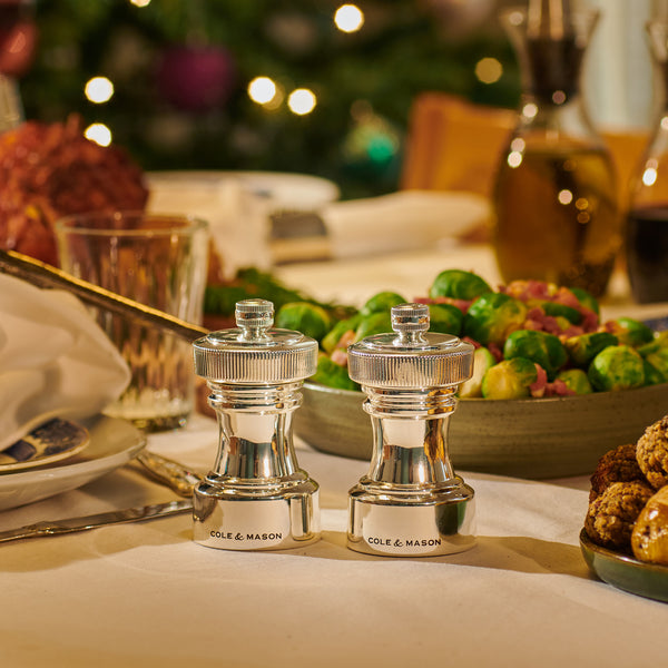 Two shiny Hoxton Silver Plated Mills by Cole & Mason sit on a festive table with holiday dishes, perfect for Christmas Gift Hampers, as blurred lights glow in the background.