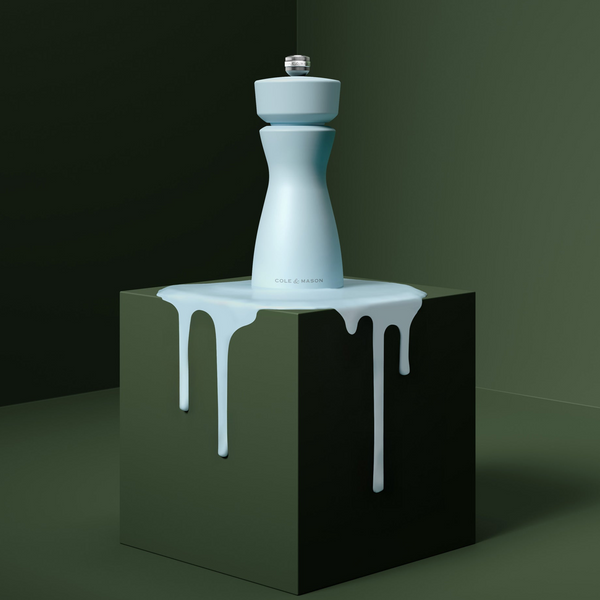 A pale blue Mix & Match Kenton Mill by Cole & Mason, featuring adjustable grind settings, stands on a dark green cube with white liquid dripping down the sides, all set against a minimalist dark green backdrop.