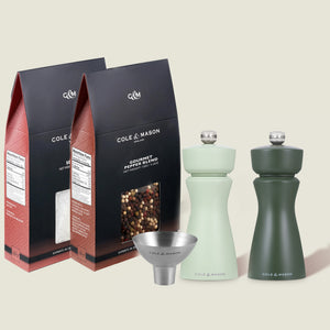 Two packets of Fast Bundle Mix & Match Kenton Mills pepper blend stand behind two green pepper mills—one light, one dark—both with adjustable grind settings, accompanied by a small stainless steel funnel.
