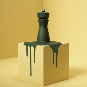 A dark green Mix & Match Kenton Mills grinder by Cole & Mason sits on a light yellow cube, with matching green liquid paint dripping down the sides, set against a minimalist yellow background.