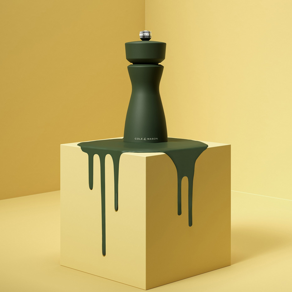 A dark green Mix & Match Kenton Mills grinder by Cole & Mason sits on a light yellow cube, with matching green liquid paint dripping down the sides, set against a minimalist yellow background.