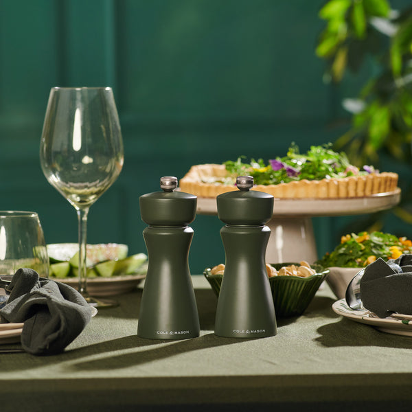 A set dining table displays the Cole & Mason Kenton Mills in Rosemary Christmas Gift Hamper, with glassware, plates and napkins, salad, and quiche arranged on a green background accented by foliage.