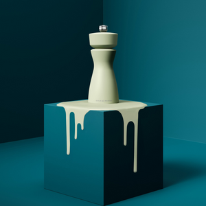 A pale green Mix & Match Kenton Mills pepper grinder by Cole & Mason sits on a teal pedestal with matching green liquid dripping down, all set against a solid teal background for a modern, minimalist look.