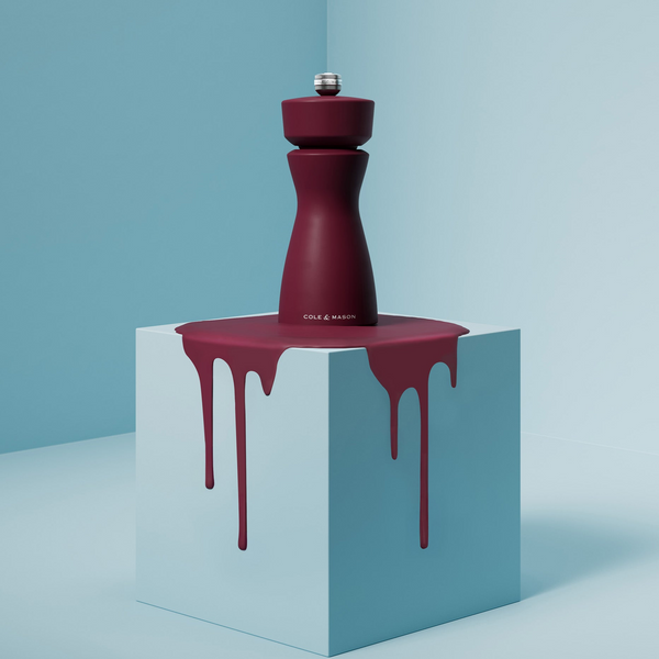 A burgundy Mix & Match Kenton Mills grinder by Cole & Mason sits on a light blue cube with burgundy liquid dripping down, creating a bold contrast against the minimal pastel blue background.
