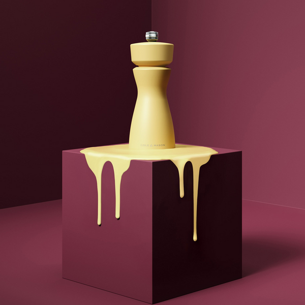 A yellow Mix & Match Kenton Mills grinder by Cole & Mason rests on a maroon pedestal, with matching yellow liquid dripping down the sides, set against a maroon backdrop.