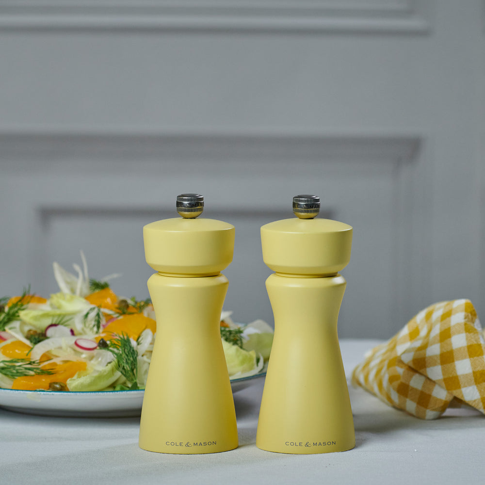 The Cole & Mason Kenton Salt & Pepper Mill in Turmeric Yellow features a silver top knob, brand logo at the base, and a durable ceramic grinding mechanism. Its photographed on a plain light background with a soft shadow to the right.