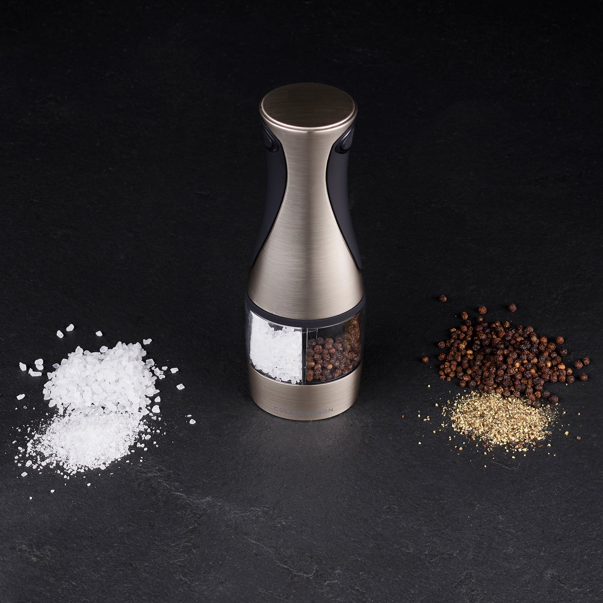 The Cole & Mason Kew Electronic 2-in-1 Duo Mill stands on a dark surface, with coarse salt on the left and whole and ground black pepper on the right, featuring adjustable grind settings for precise seasoning.