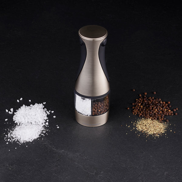 The Cole & Mason Kew Electronic 2-in-1 Duo Mill stands on a dark surface, with coarse salt on the left and whole and ground black pepper on the right, featuring adjustable grind settings for precise seasoning.