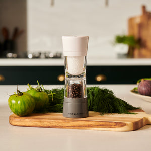 The Cole & Mason Lincoln 2-in-1 Duo Mill with adjustable settings rests on a wooden cutting board beside fresh green tomatoes and dill, set in a modern kitchen with blurred background elements.