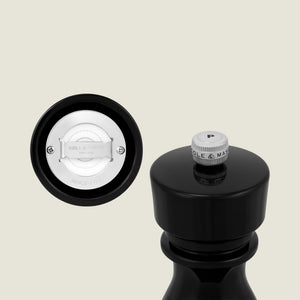 A close-up of the Cole & Mason London Black Acrylic Pepper Mill 150mm, topped with a silver 'P' knob, sits beside its white premium grinder mechanism against a light beige background.
