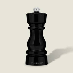 The Cole & Mason London Black Acrylic Salt & Pepper Mills 150mm features a glossy hourglass design and premium grinding technology, shown standing upright against a light beige background.