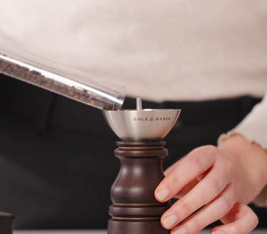 A person pours peppercorns into a Cole & Mason London Chocolate Wood Mill from the Christmas Gift Hamper, holding the mill steady as part of their festive preparation.
