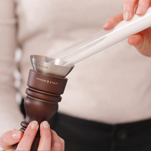A person in a light sweater with manicured nails pours salt into a pepper grinder using a metal funnel labeled Cole & Mason—from the London Chocolate Wood Mill & Shaker Christmas Gift Hamper.