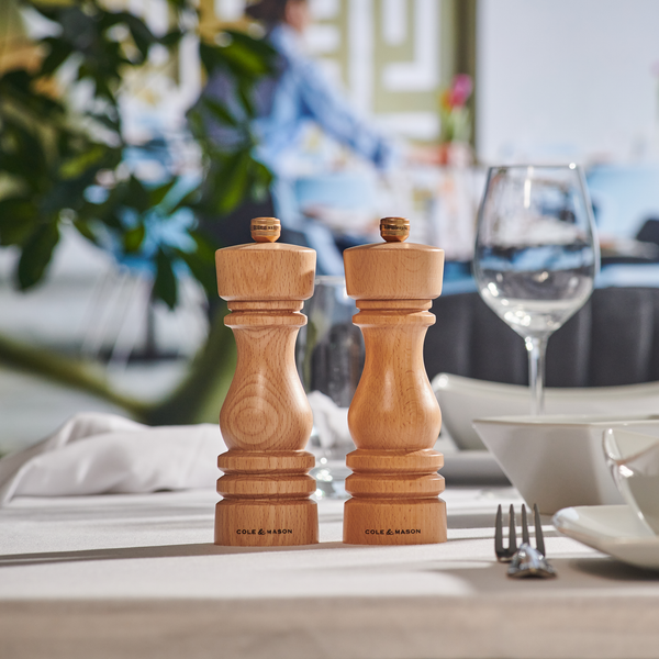 A Cole & Mason London Natural Beech Mill Gift Hamper sits on a set dining table with dishes, glass, and cutlery in a bright restaurant. The softly blurred background makes it ideal for gift hampers or elegant dining settings.