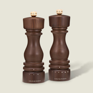 Two Cole & Mason London Walnut Salt or Pepper Mills stand side by side on a neutral background. The pepper mill is positioned on the left, while the adjustable salt grinder sits on the right; each features distinct grind size indicators at the base.