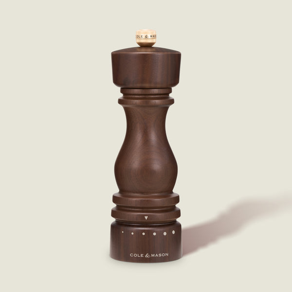The Cole & Mason London Walnut Salt or Pepper Mill, featuring a rounded dark wood walnut design and labeled with the brand name at the base, stands upright on a light beige background. Adjustment dots and a small knob are visible at the top and base of this elegant seasoning mill.