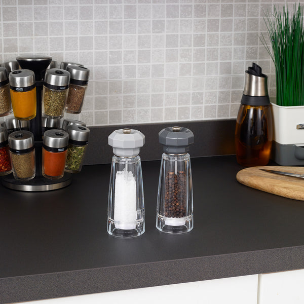 The Cole & Mason UK Lowestoft Salt & Pepper Mill Set 175mm, with clear acrylic bodies and adjustable ceramic grinders, sits on a black kitchen counter alongside a spice rack, knife block, plant, and wooden cutting board.