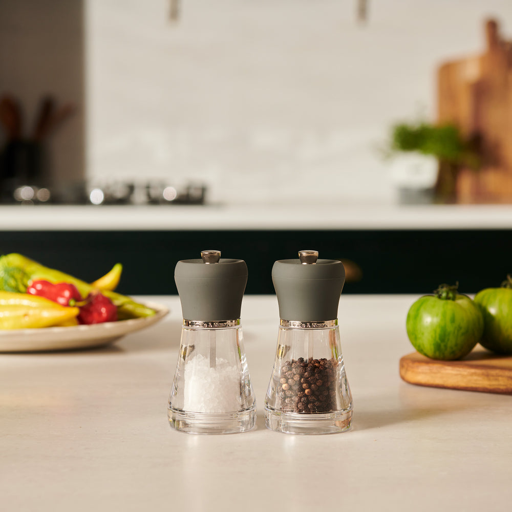 The Cole & Mason Napoli Salt & Pepper Mill Set features elegant grinders with gray tops, high-quality mechanisms, and comes filled with coarse salt and whole black peppercorns on a light background.