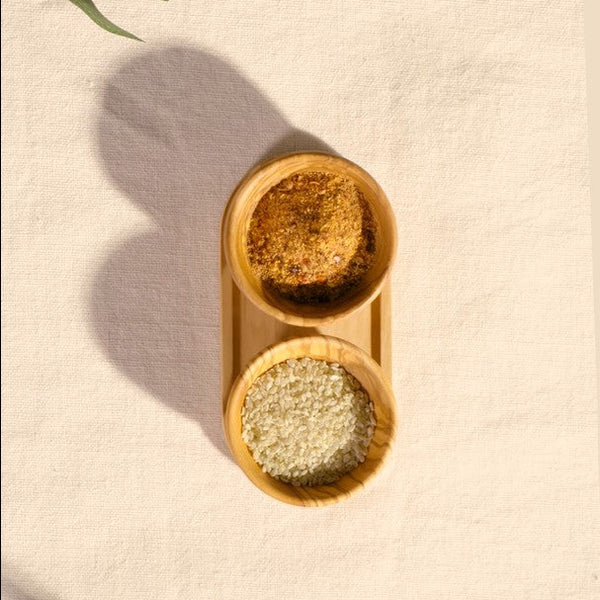 The Cole & Mason Portobello Olive Wood Pinch Pot Set features two small bowls, one filled with a brown spice blend and the other with white grains, their fine olive wood patterns shown on a textured surface.