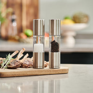 The Cole & Mason Oslo Salt & Pepper Mill Set, featuring adjustable grinding mechanisms, sits on a kitchen counter beside cooked meat on a wooden board. A softly blurred background highlights precise seasoning control.