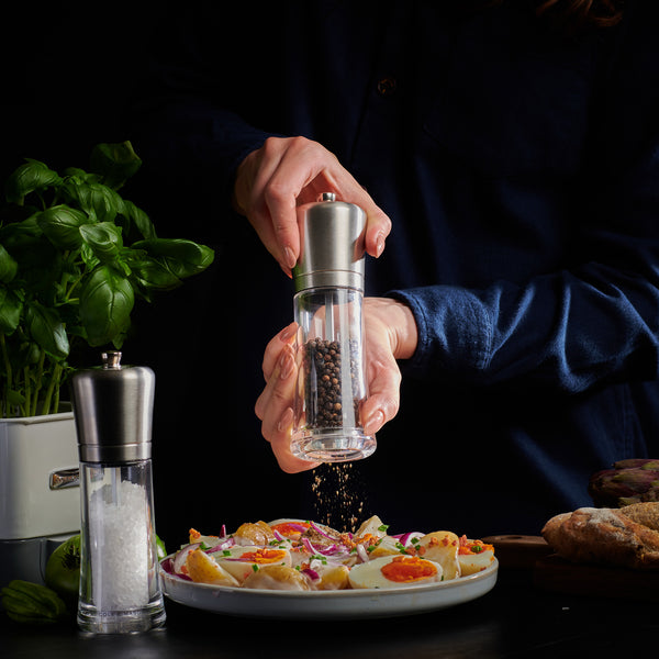 Someone grinds black pepper with the Cole & Mason Sandown Salt & Pepper Mill Set, featuring adjustable grinding for precise seasoning. A salt grinder, fresh basil, and bread sit nearby on the table against a dark backdrop.