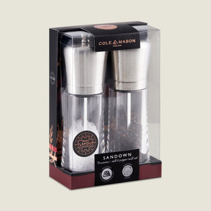 The Cole & Mason Sandown Salt & Pepper Mill Set features stainless steel tops and clear bodies for seasoning control, displayed in mostly black packaging with gold and red accents, showing salt and peppercorns in each mill.
