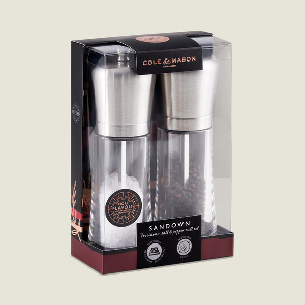 The Cole & Mason Sandown Salt & Pepper Mill Set features stainless steel tops and clear bodies for seasoning control, displayed in mostly black packaging with gold and red accents, showing salt and peppercorns in each mill.
