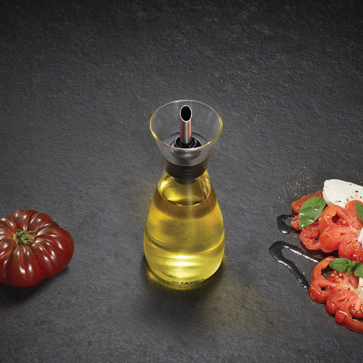 A glass oil dispenser filled with yellow olive oil sits on a dark stone surface, with a heirloom tomato on the left and a sliced tomato salad with mozzarella, basil, and olive oil on the right.