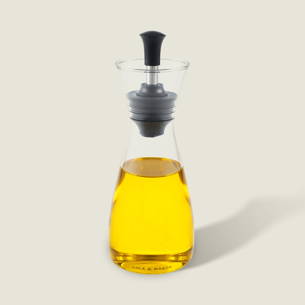 The Cole & Mason Sawston Oil & Vinegar Pourer features yellow oil inside, an ergonomic glass carafe with a black and gray stopper, and a labeled base. It casts a soft shadow on a light background.