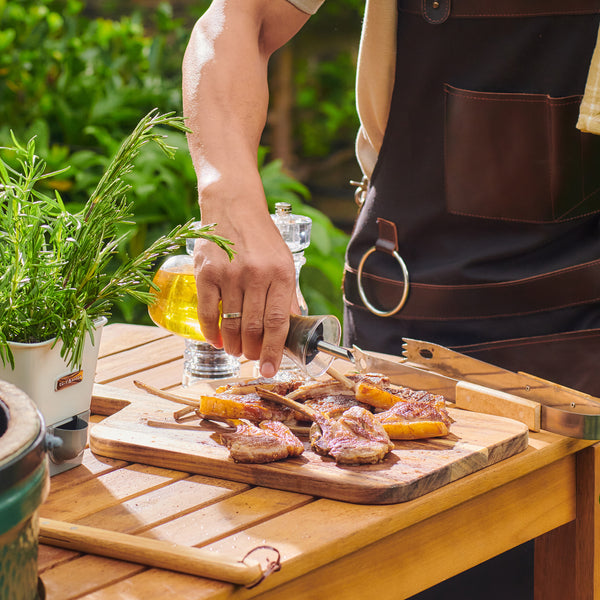 Wearing an apron, a person uses the Cole & Mason Sawston Oil & Vinegar Pourer to drizzle beer over grilled skewers on a wooden board outdoors, with fresh herbs and greenery in the background.