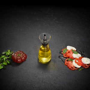 The Cole & Mason Sawston Oil & Vinegar Pourer, with its ergonomic design, sits on a dark surface between fresh tomato and basil and a tomato, mozzarella, and basil salad—ideal for controlled pouring onto any dish.