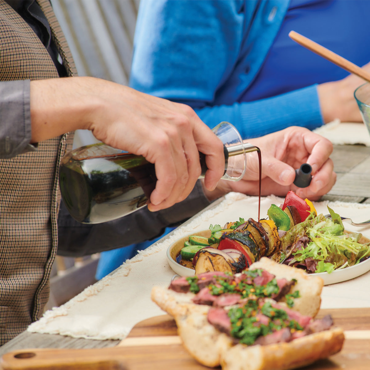 A person pours balsamic dressing onto a colorful salad beside grilled vegetables and sliced steak on bread, at an outdoor table. Another person in blue clothing sits nearby.