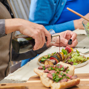 A person pours dressing from the Cole & Mason Sawston Oil & Vinegar Pourer onto a colorful vegetable skewer by a fresh salad, while a steak and herb sandwich sits up front. Another person is seated across the table.