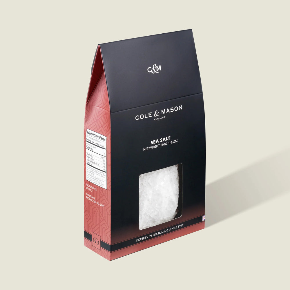 The Cole & Mason Sea Salt Box features a black and red design with a clear window revealing coarse sea salt crystals inside. Ideal for salt mills, the packaging includes product details and a nutrition facts label on the side.