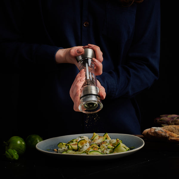 #pepper
A person in a dark shirt uses the Cole & Mason UK Windermere Pepper Mill 165mm to season sliced zucchini, with green tomatoes and bread on the table. A dark background highlights the scene.