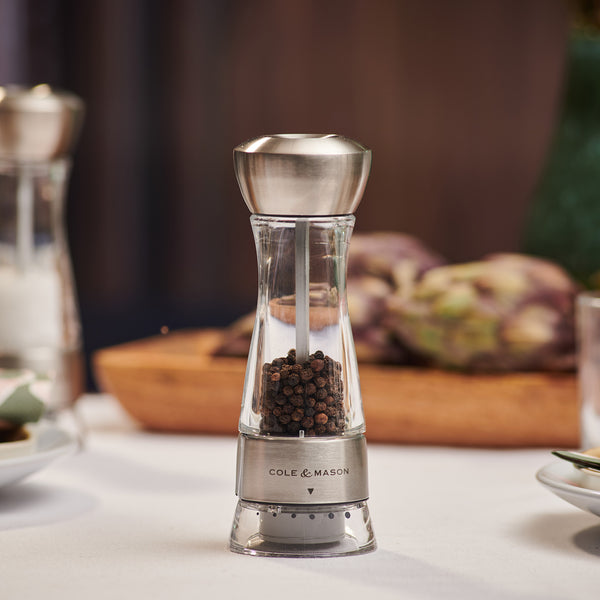 #pepper
The Cole & Mason UK Windermere Pepper Mill 165mm, made of clear acrylic and stainless steel and filled with black peppercorns, sits on a dining table. The Cole & Mason UK branding is visible on the metal band. Blurred food and tableware are in the background.