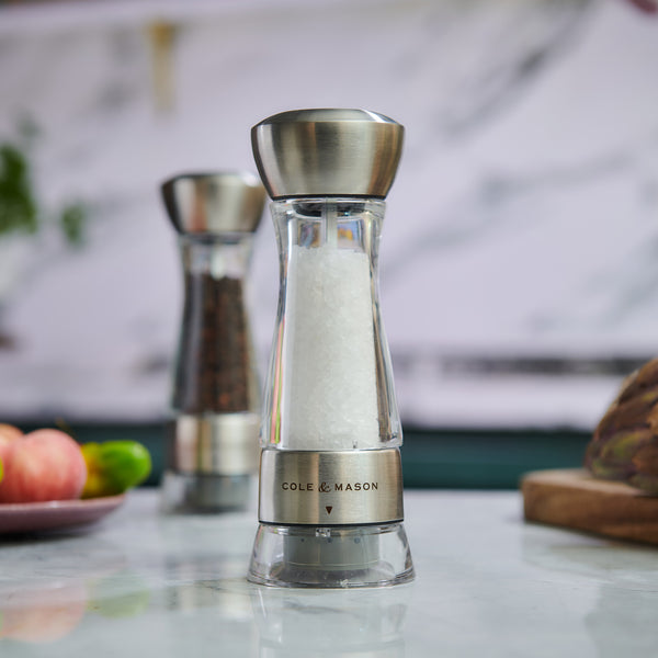 #salt
A close-up of the Cole & Mason Windermere Pepper Mill 165mm, crafted from crystal clear acrylic and filled with peppercorns, showcases its adjustable grind precision. A matching salt grinder and assorted vegetables appear blurred in the background.