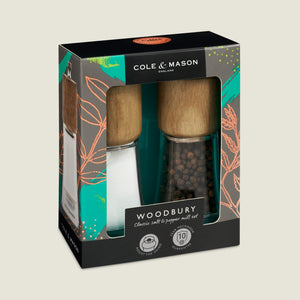 The Cole & Mason Woodbury Salt & Pepper Mill Set features natural wood tops, adjustable coarseness, and comes pre-filled—one with salt, one with black peppercorns. Box showcases a teal and orange leafy design.